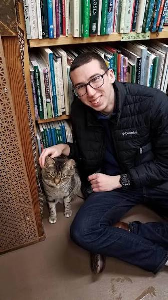 Alex Kearns smiling beside a cat in front of a bookshelf